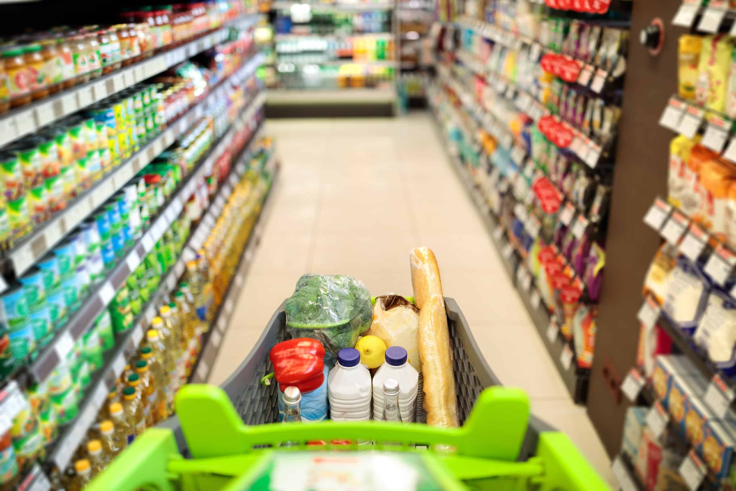 Shopping Cart Full Of Food Products Over Blurred Supermarket Aisle Background. POV Of Customer Pushing Trolley Walking In Grocery Store Along Shop Shelves. Selective Focus